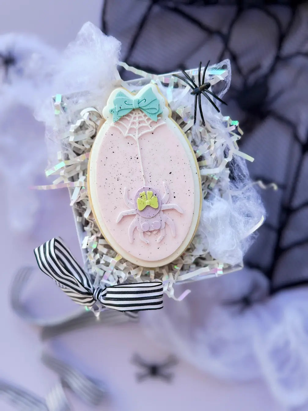 Ovaler hellrosa Fondantkeks mit Spinne, Spinnennetz und türkisfarbener Schleife.