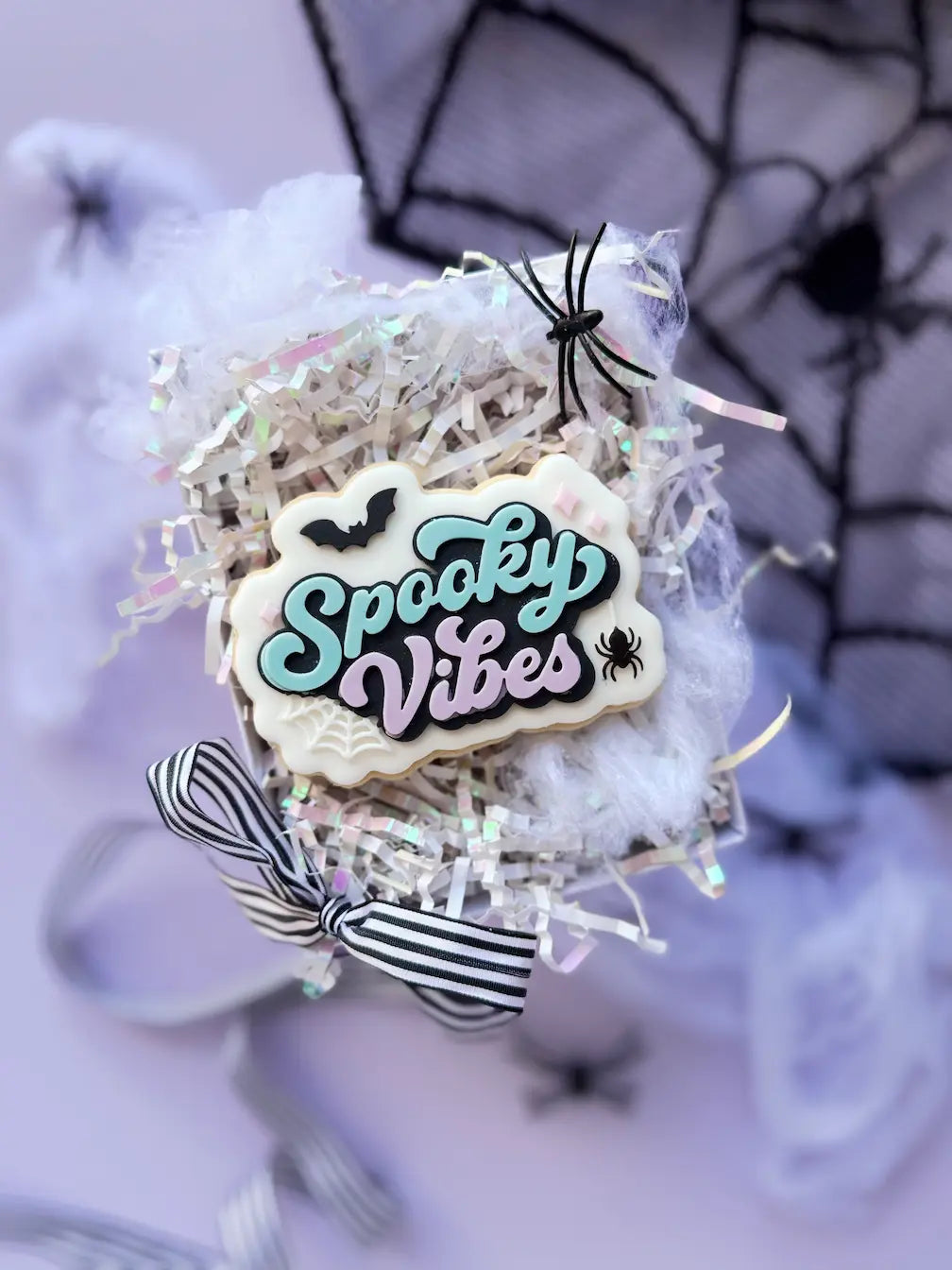 Dekorierter Keks mit Spooky Vibes Schriftzug in Türkis und Flieder, schwarzer Fledermaus, Spinne und weißem Spinnennetz.