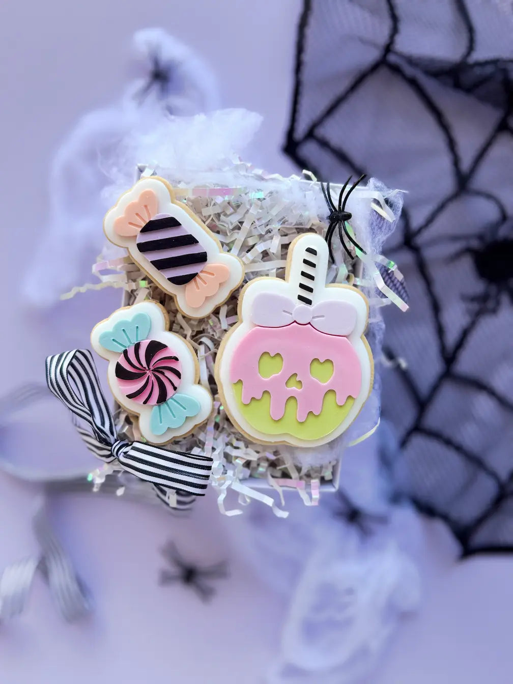 Drei dekorierte Halloween-Kekse: zwei Bonbons und ein grüner Apfel mit rosa Tropfenglasur in Totenkopfform und Schleife.