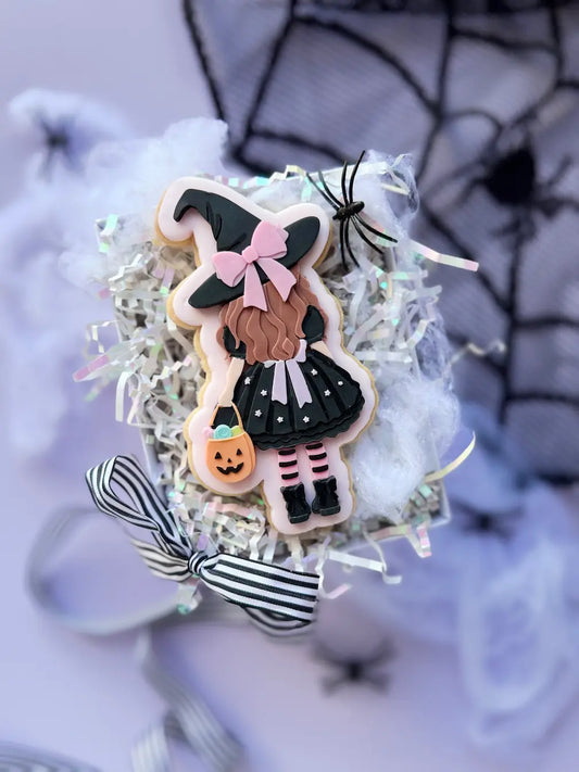 Dekorierter Halloween-Keks als Mädchen von hinten mit schwarzem Hexenhut, rosa Schleife und Kürbistasche.