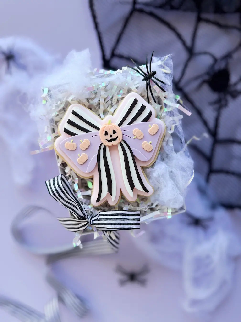 Dekorierter Halloween-Keks als große Schleife mit schwarzen Streifen, fliederfarbenen Schleifenflügeln und kleinen Kürbissen.