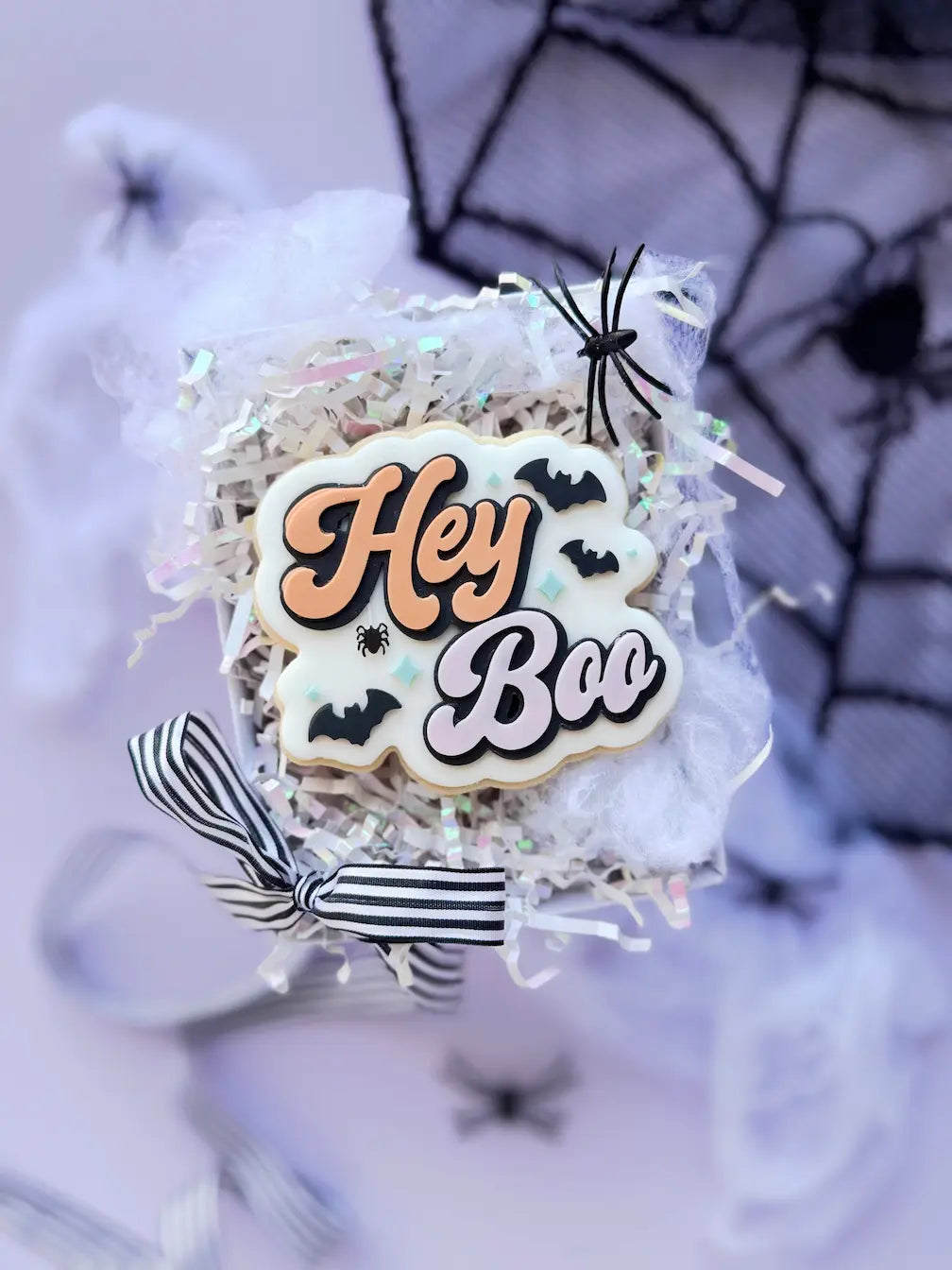 Dekorierter Halloween-Keks mit Hey Boo Schriftzug in Orange und Flieder, schwarzen Fledermäusen und kleiner Spinne.
