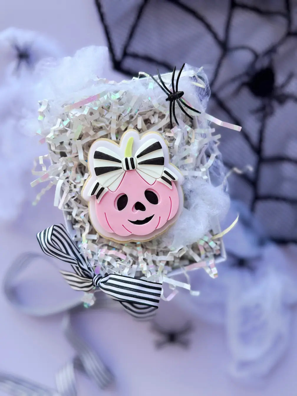 Dekorierter Halloween-Keks als rosa Kürbis mit lächelndem Gesicht und großer schwarz-weiß gestreifter Schleife.