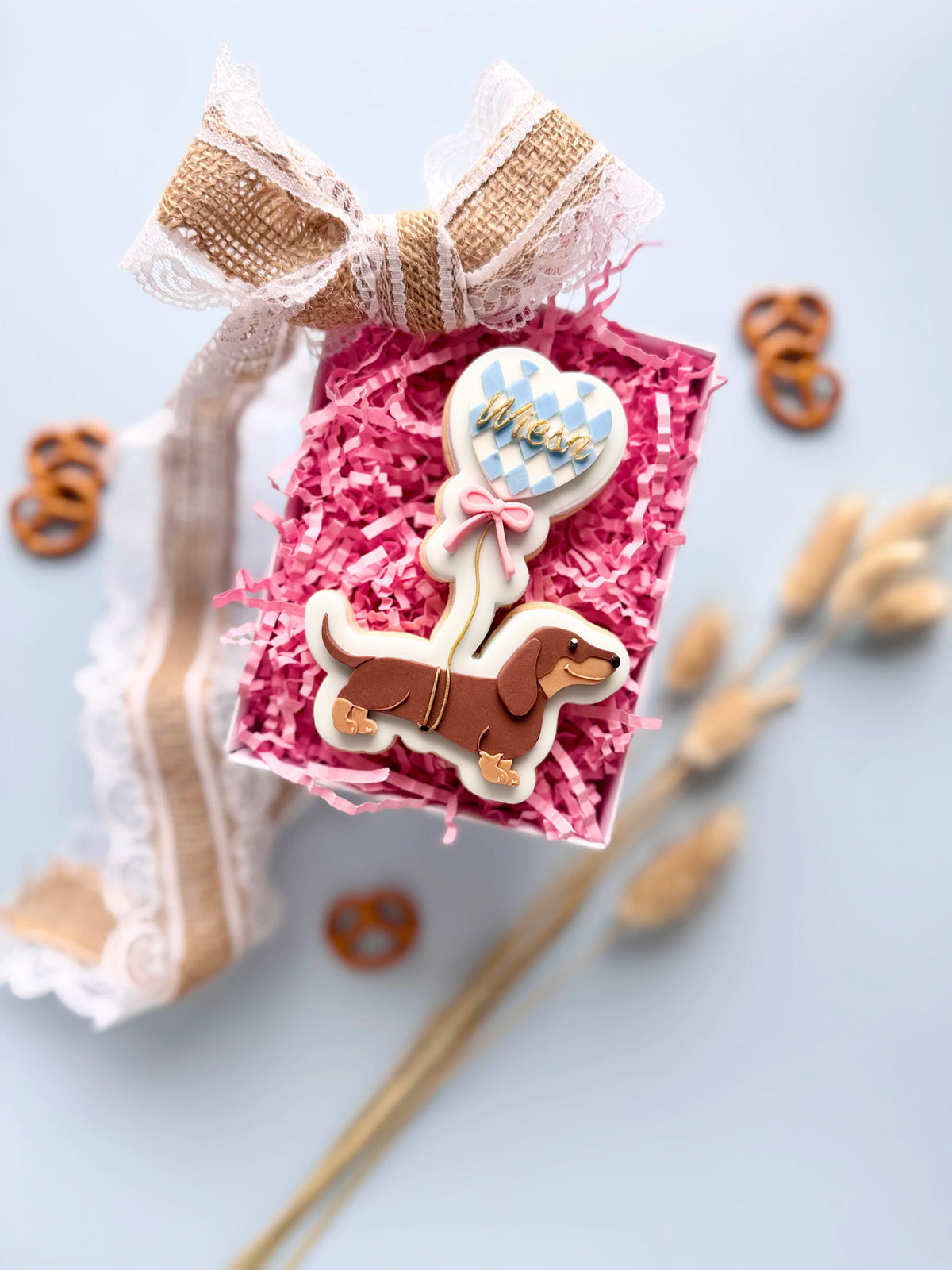 Verzierter Oktoberfest-Keks mit braunem Dackel und blau-weißem Herzballon in einer Geschenkbox mit pinkem Füllpapier.